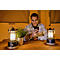 Un jeune homme en chemise à carreaux est assis à une table, souriant et regardant son smartphone, entouré de deux lanternes et d'une petite plante.