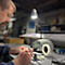 Un homme travaille à un bureau sous une lampe, tenant des outils et travaillant sur une petite pièce métallique.