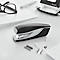 Black stapler with white accents, pens, glasses, hole punch, and paper on a white table. LEITZ logo.