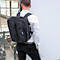 Un homme avec un sac à dos noir se tient contre un mur blanc. Il porte une chemise blanche et un pantalon noir.