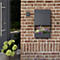 Boîte aux lettres murale gris foncé avec jardinière sur mur de briques. A côté, porte d'entrée et panier avec fleurs. Sonnette au-dessus de la boîte.