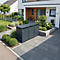 Trois poubelles grises devant une maison moderne. Sentier pavé, jardin avec buissons et arbres.