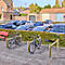 Zwei Fahrräder stehen in einem hölzernen Fahrradständer auf einem gepflasterten Platz vor einer Hecke und einem Parkplatz.
