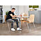Man sitting at table, writing. Wooden chairs with metal legs. Light wood, grey trousers, black t-shirt, white sneakers.