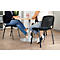 Zwei Personen sitzen an einem runden Tisch, beide tragen Jeans und Turnschuhe. Der Hintergrund ist ein Büro.