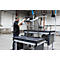 Een man bedient een robotarm die een metalen mand op een tafel inspecteert. Fabriek op de achtergrond.