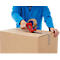 Person sealing a cardboard box with tape. Bright clothing and a red tape gun are visible.