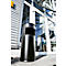 Black trash can in front of glass facade. The bin has a rectangular opening. Buildings and sky can be seen in the background.
