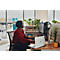 A woman sits at a desk in front of a computer. She is wearing a red top and has dark hair. There is a laptop and potted plants on the desk.