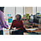 A woman in a red blouse is seated, looking up at a person standing in front of her, holding a book. A desk with computers is visible.