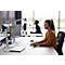 A woman wearing a headset works at a computer in an office. A man in the background, also wearing a headset.