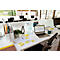 Bureau met laptop, notities, pennen, groene mok en plant. Andere werkplekken op de achtergrond.