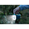 Hand holding a glowing, blue flashlight in a forest. The beam of light illuminates the path.