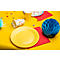 Assiette et gobelet jaunes, boule en papier bleue, serviettes rouges sur table jaune, bonbons, décoration de fête.