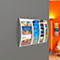 White curved magazine rack with brochures on grey wall. In the background, an orange wall section with floor lamp.