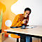 A smiling woman drinks tea at a desk. She is wearing an orange top and sitting at a standing desk.