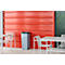 Grijze vuilnisbak voor rode muur, naast witte tafels.