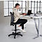 Un homme est assis à un bureau réglable en hauteur, travaillant sur un ordinateur portable. Il est assis sur une chaise pivotante noire avec repose-pieds. Le bureau est lumineux.
