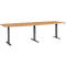 A long, rectangular table with a light brown tabletop and gray-black, height-adjustable legs. The table is shown in detail.