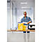 A man with glasses stands smiling at a table with a printer and paper, holding paper in his hand. Yellow paper stacks next to it.
