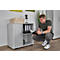Man squats by a gray cabinet, reading a book. The cabinet has a roller shutter. File folders sit on the shelf.
