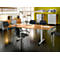 A conference room featuring a light wood table, black chairs, and windows. A long light bar hangs above the table.