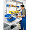 A man in blue workwear sorts parts at an industrial workstation. Shelves with containers and lighting.