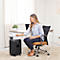 A woman sits at a desk placing documents into a paper shredder. She wears a blue blouse and dark trousers.