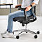 Personne assise sur une chaise de bureau. Dossier en maille bleu clair, assise noire, cadre argenté, roulettes.