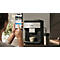 Person bedient Kaffeemaschine via Smartphone. Auf dem Tisch stehen Kaffeemaschine, Tasse, Thermoskanne.