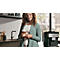 Frau mit lockigem Haar hält Latte Macchiato, lächelnd in der Küche. Kaffeemaschine im Hintergrund.