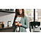 Eine Frau mit lockigem Haar hält einen Latte Macchiato in der Küche. Im Hintergrund eine Kaffeemaschine.