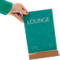 A hand holds a table card with the word LOUNGE, standing on a wooden base. Green lines and text in the background.