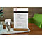 Menu card on a wooden stand. Various cutlery in a glass, white napkin paper and a green chair in the background.