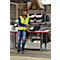 Een man in een gele vest leunt op een tafel in een magazijn en glimlacht naar de camera. Achter hem staan planken en computers.