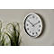 A round silver wall clock with black markings hangs on a light grey wall. Green plants are visible behind it.