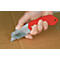 A knife is cutting cardboard. The handle is red, the metal is silver. The hand is holding the knife.