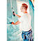 A man fastens foil to a wall with a stapler. He is wearing jeans and a white t-shirt.