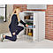 A woman kneels in front of a white cabinet, opens it, and reaches for an item.