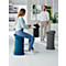 A woman sits on a blue ergonomic stool while a man stands in an office. Two other stools are visible.