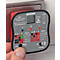Een rode stopcontacttester met grijze bedieningselementen wordt door een hand voor een stopcontact gehouden. De tester toont verschillende resultaten.