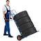 A man in overalls pushes a blue hand truck loaded with five car tires. He is smiling.