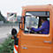 A man driving an orange truck. A convex road traffic mirror is visible through the windshield.