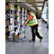 A man in a warehouse pushes a loaded trolley. He wears a yellow safety vest and stands in front of warehouse shelves.