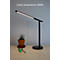 Schlanke, schwarze Schreibtischlampe mit leuchtendem LED-Arm auf einem Holztisch, daneben eine Topfpflanze und Stifte.