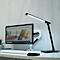 Schreibtisch mit Computer und schwarzer LED-Schreibtischlampe, die leuchtet. Auf dem Monitor ist eine Webseite zu sehen.