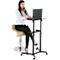 A woman sits at a height-adjustable standing desk with a laptop. She wears a white shirt and black trousers.