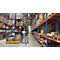 Man with a laptop in a warehouse between racks of boxes. Blue and orange metal shelving.