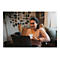Eine lÃ¤chelnde Frau sitzt vor einem Laptop und hÃ¤lt eine Tasse. Im Hintergrund sind Fotos an der Wand und eine Topfpflanze zu sehen.