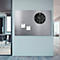 Zwarte wandklok met plaknotities op een metalen plaat tegen een lichtblauwe achtergrond. In de verte is een kantoorgang te zien.